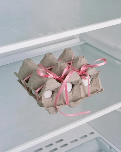 Egg carton with pink ribbon inside a refrigerator. The carton holds a dozen white eggs, displayed in a clean, bright refrigerator setting.