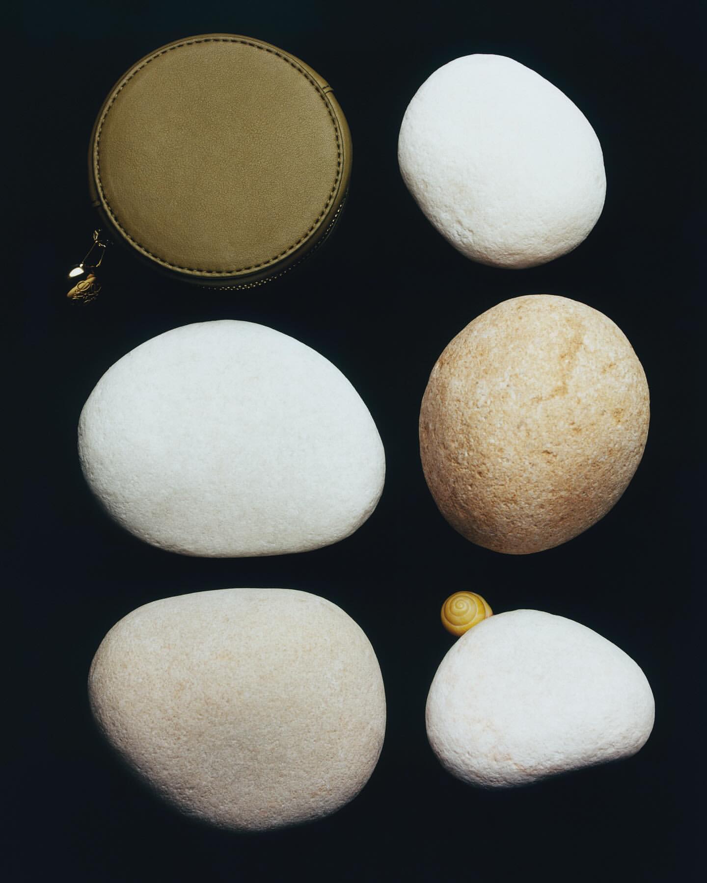 Overhead still life: A round, olive-green zippered case and five smooth, light-colored stones are set against a dark backdrop.