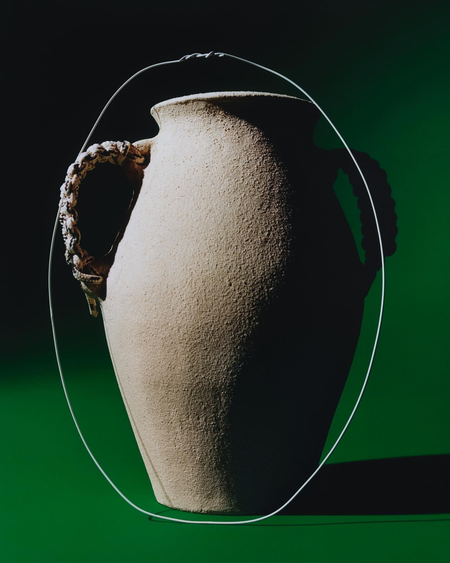 Tan ceramic vase on a green surface features a slender metal wire and decorative rope-like handles. The light source is above.