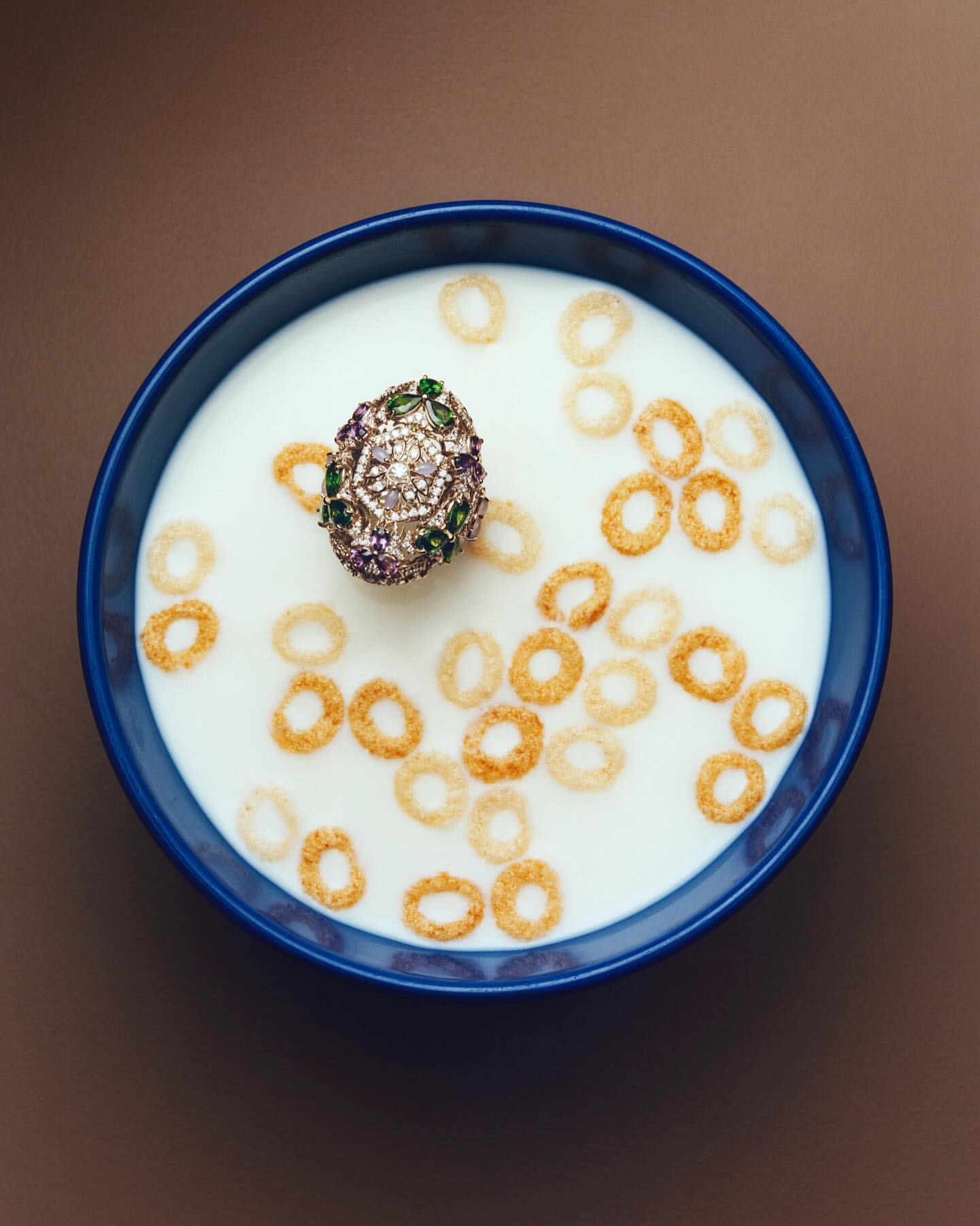 Overhead studio shot of an embellished ring in a blue bowl of cereal and milk on a brown background showcasing food and jewelry photography.