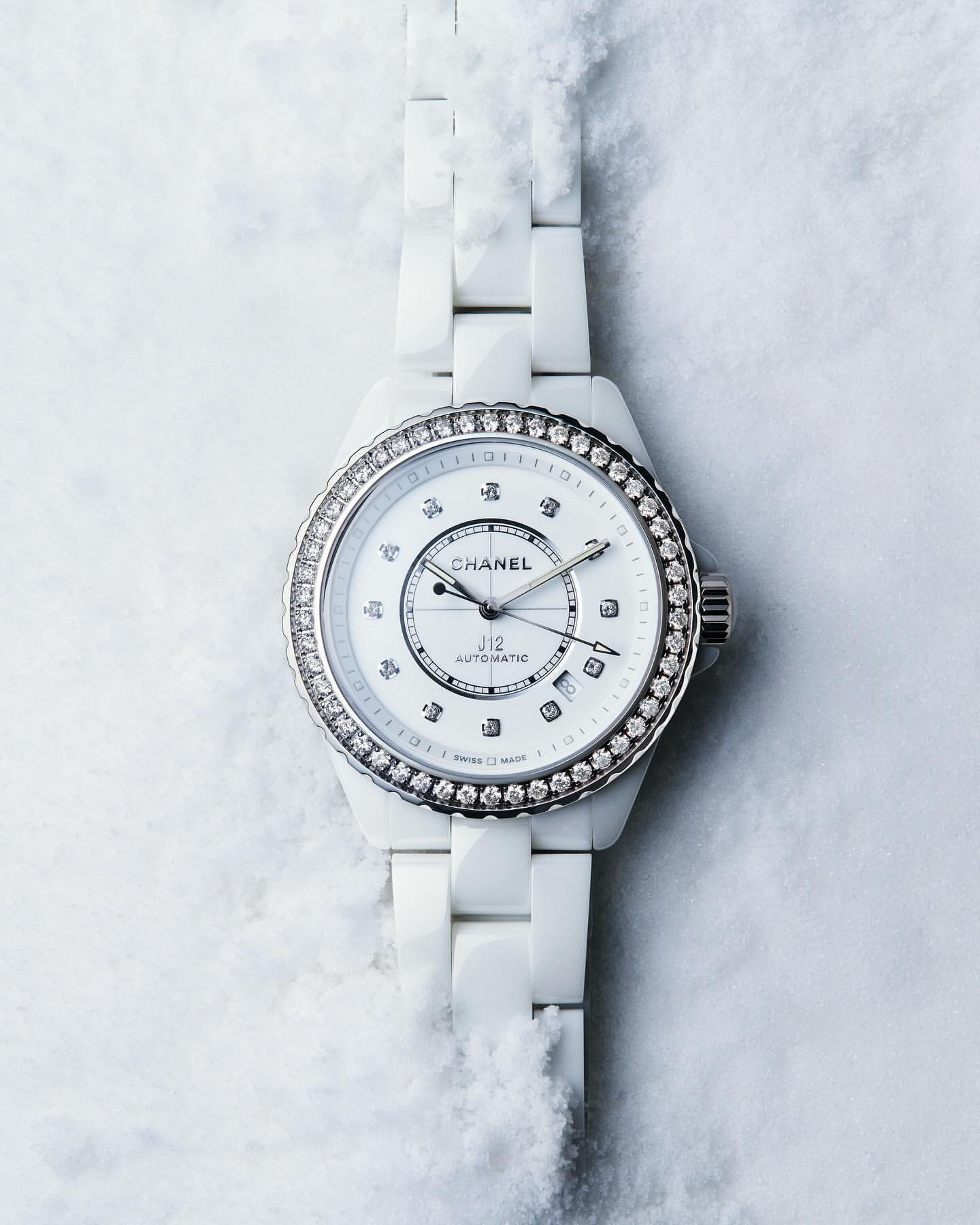 Chanel J12 watch close-up on a textured white background. Wristwatch features a white ceramic bracelet and a diamond bezel.