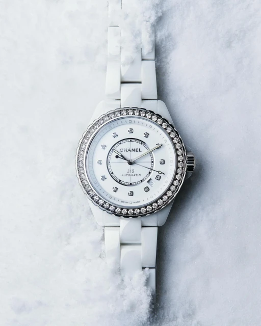 Chanel J12 watch close-up on a textured white background. Wristwatch features a white ceramic bracelet and a diamond bezel.