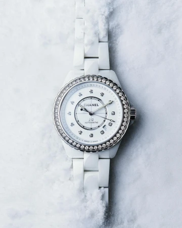 Chanel J12 watch close-up on a textured white background. Wristwatch features a white ceramic bracelet and a diamond bezel.