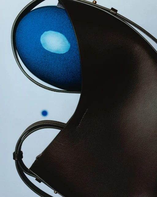Abstract close-up of a black leather tote bag with metal handles overlapping a blue reflective orb on a light blue background.