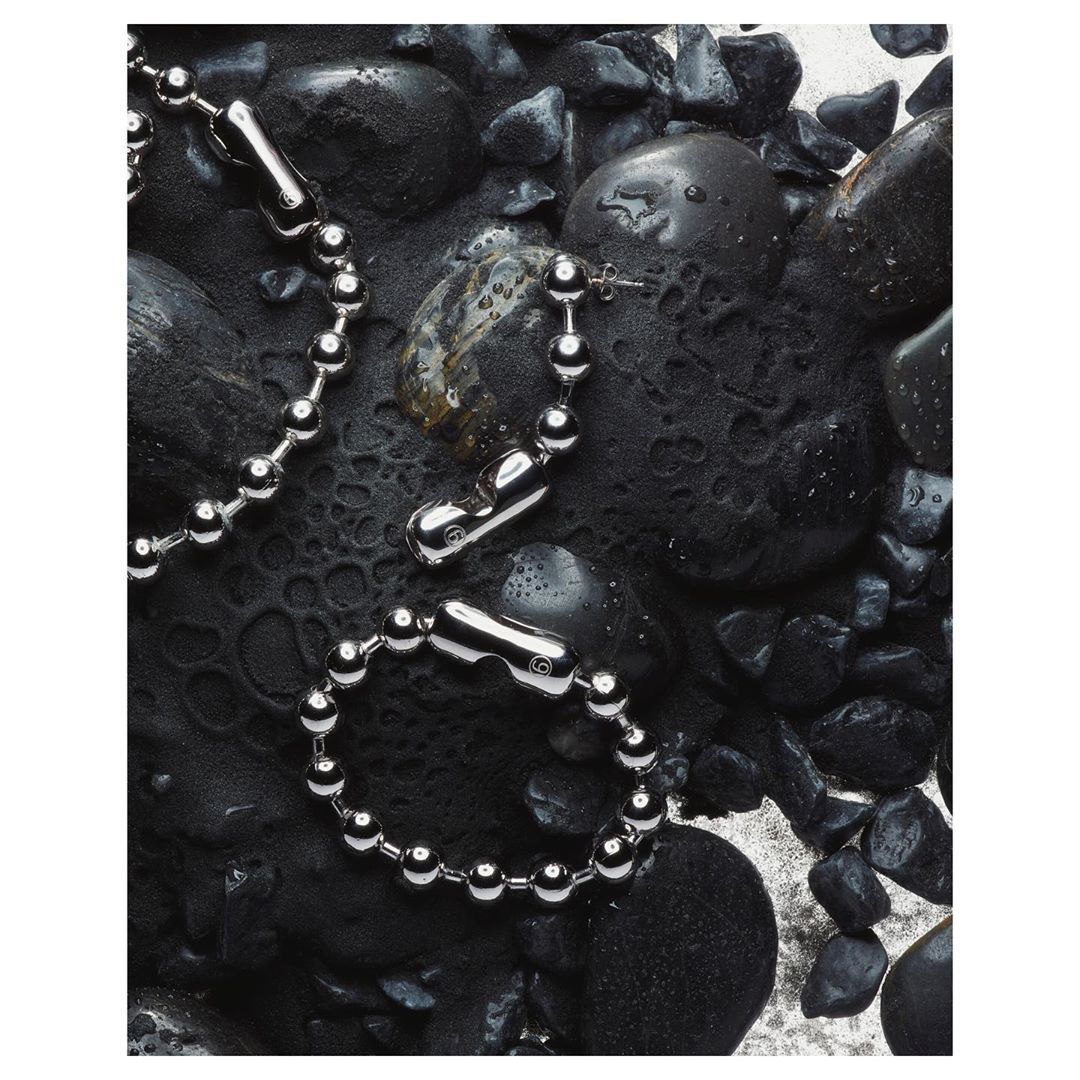 Silver chain necklace and bracelet with barbell charms on black stones and light sand background in a studio setting.