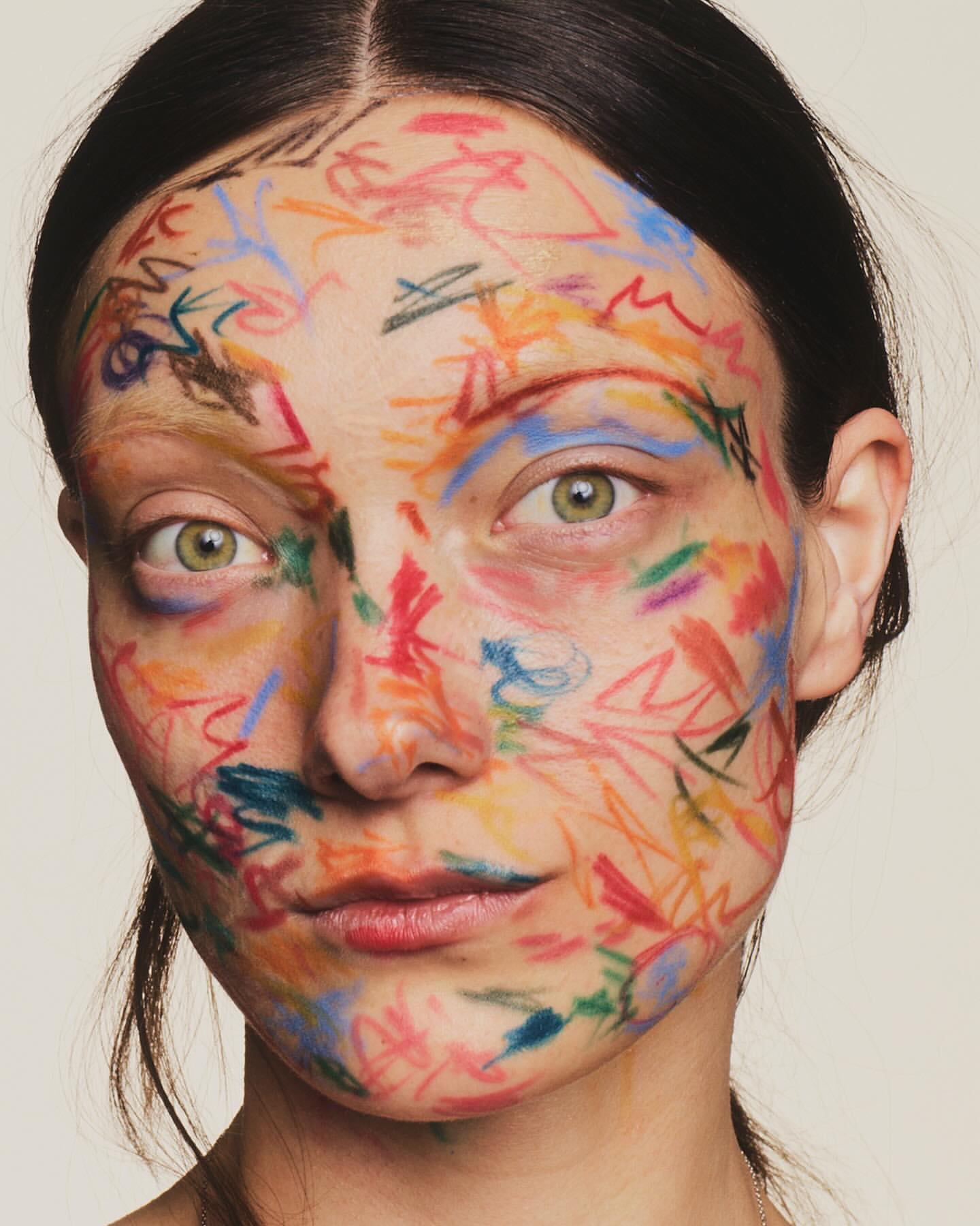 Portrait of a young woman with colorful crayon-like face paint, showcasing a unique beauty style in a close-up shot on a neutral backdrop.