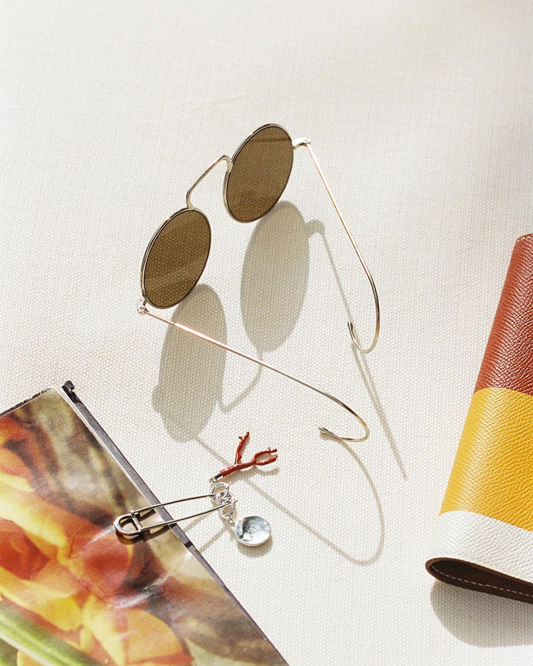 Overhead shot of round sunglasses with a gold frame, alongside a colorful wallet, a magazine page, and a decorative pin on a white surface.