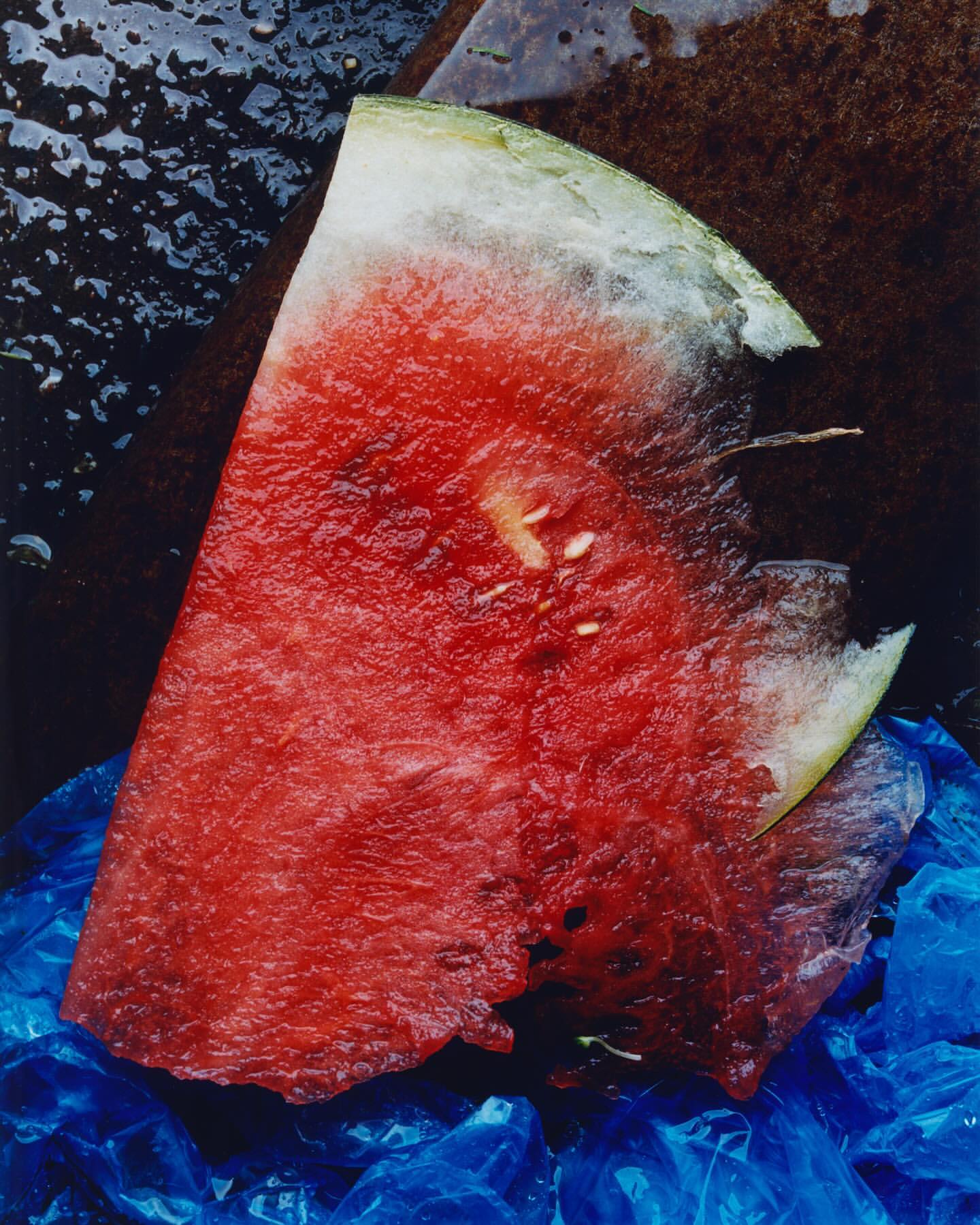 Close-up of a vibrant watermelon slice displaying its bright red flesh, scattered seeds, and a portion of rind against a plastic bag.