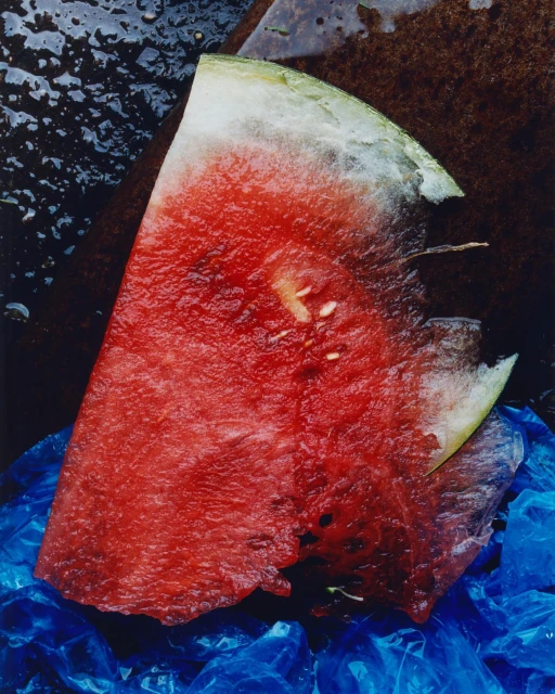 Close-up of a vibrant watermelon slice displaying its bright red flesh, scattered seeds, and a portion of rind against a plastic bag.