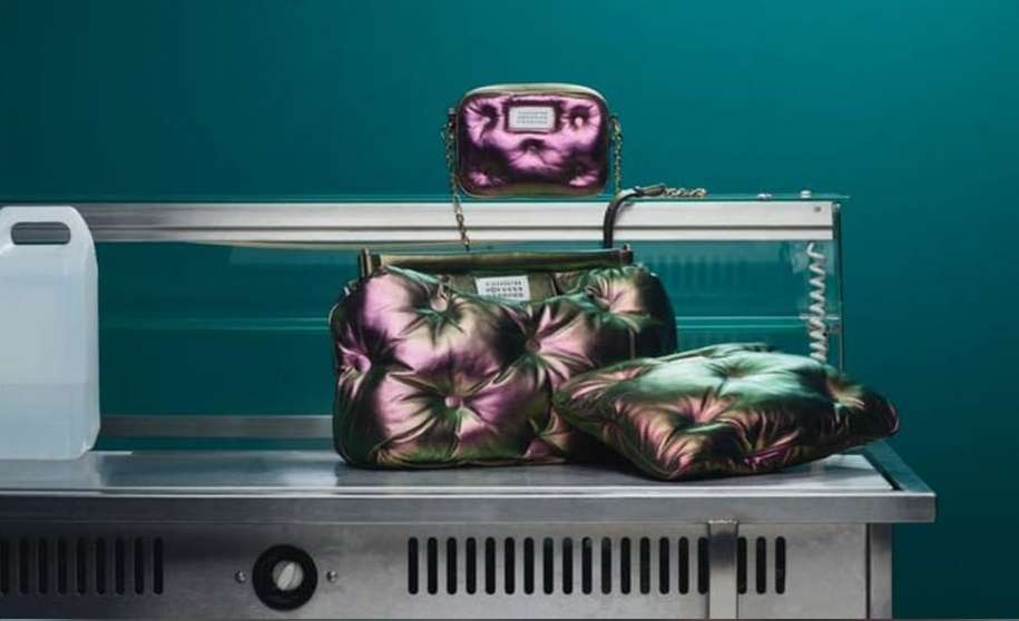 Fashion product shot of three metallic purple handbags on a stainless steel countertop against a teal wall in a studio setting.
