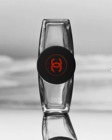 Clear glass perfume bottle with a black cap featuring a red Chanel logo, set against a soft grey and white blurred background.