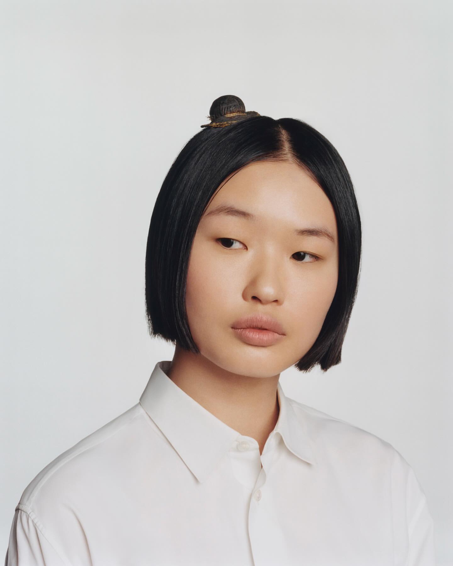 Headshot of an Asian woman with short black hair and a white button-down shirt, wearing a miniature hat on top of her head in a studio setting.
