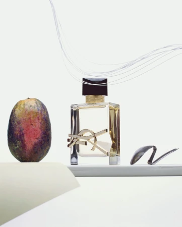 Still life shot of YSL perfume bottle with gold logo next to a mango on a white shelf against a white background with fibers.