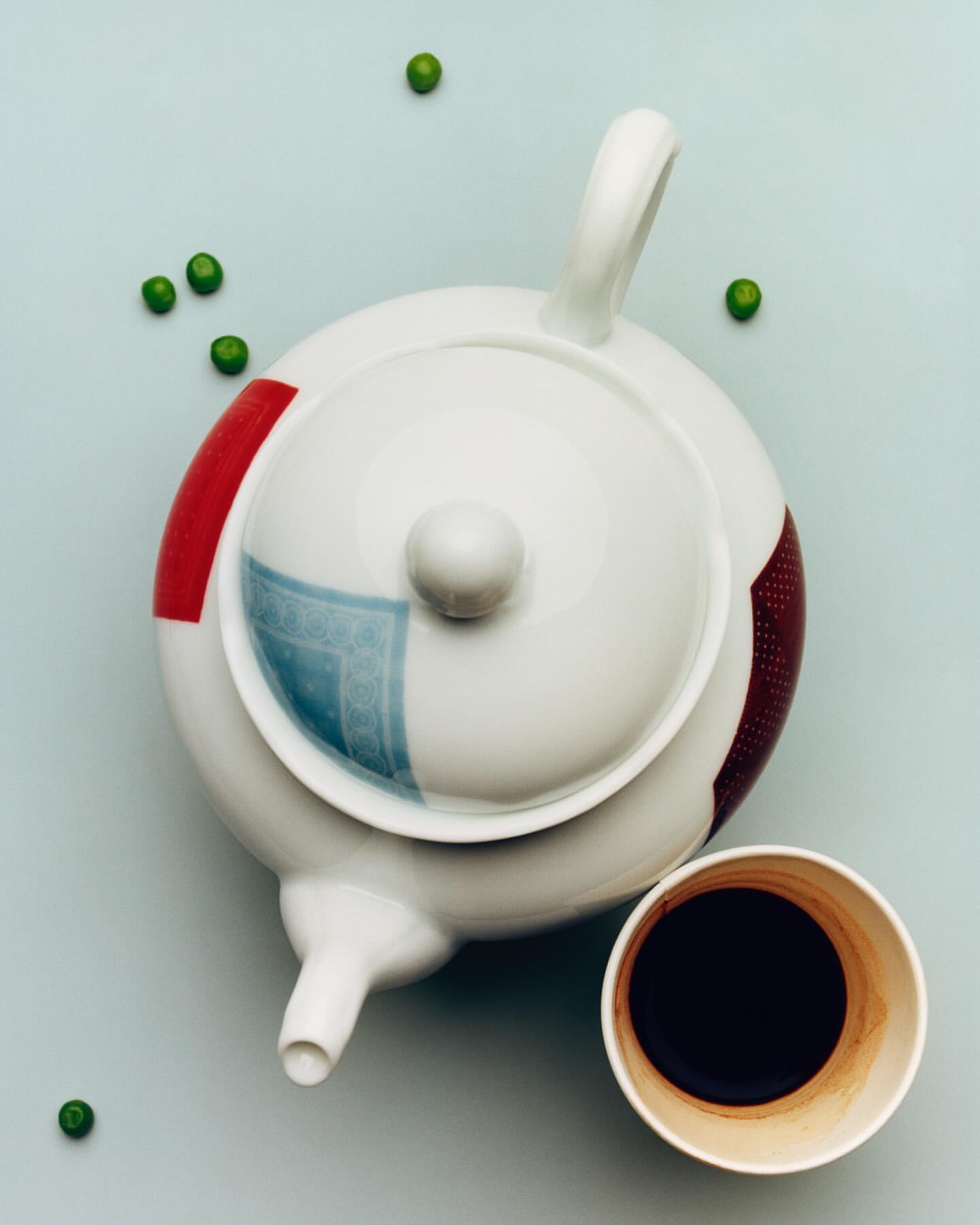 Overhead shot of white ceramic teapot with blue, red, and brown patterns alongside a small cup of dark liquid, on a light blue background.