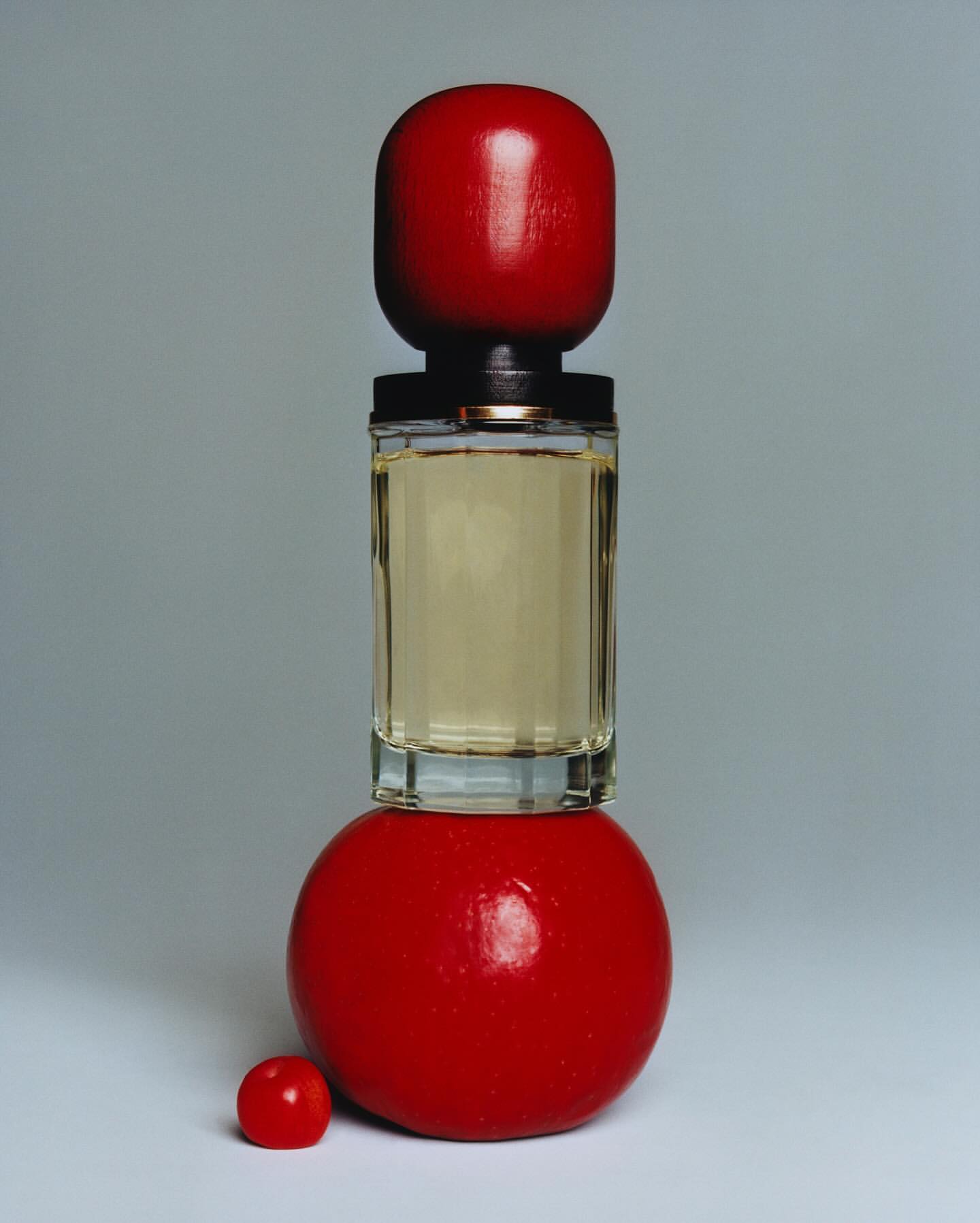 Still life of a clear glass perfume bottle with red spherical cap and base against a light gray background in studio lighting.