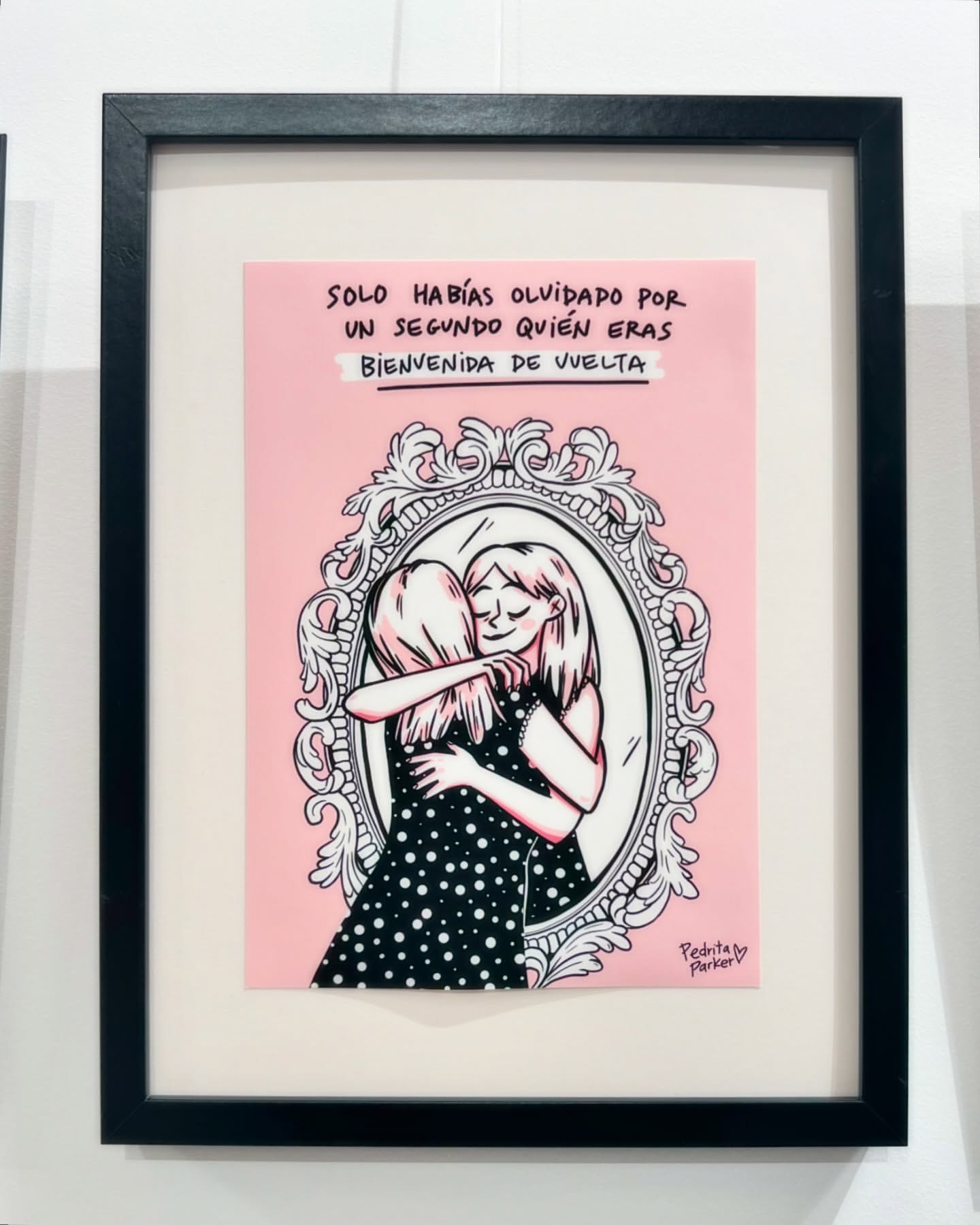 Framed art print of two cartoon women hugging in front of a mirror, on a white wall, with a message written above.