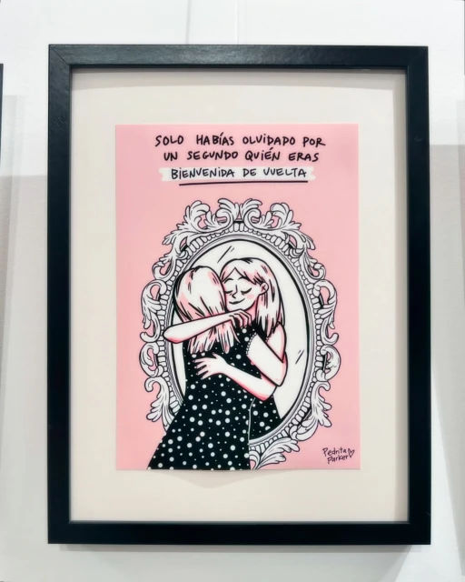 Framed art print of two cartoon women hugging in front of a mirror, on a white wall, with a message written above.