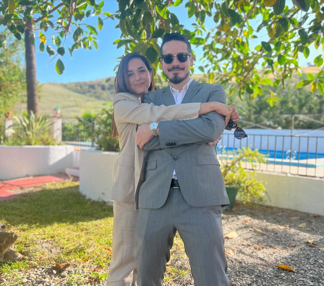 A stylish couple in suits poses outdoors. The woman wears a beige suit, and the man sports a gray suit with sunglasses and a watch.