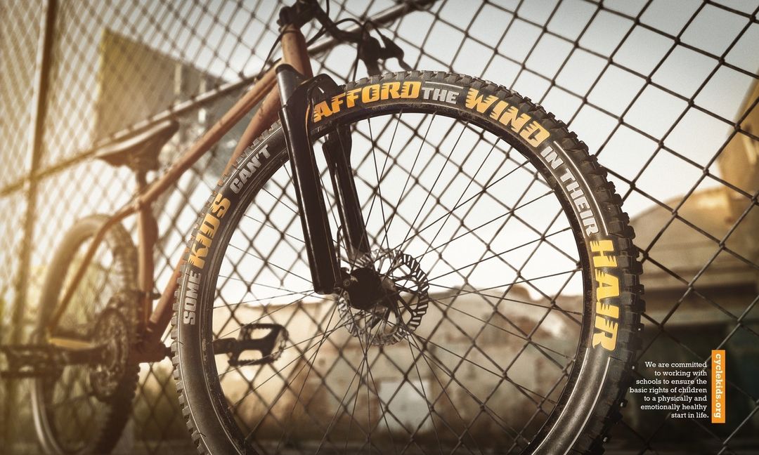 A bicycle is secured to a chain link fence with the words 'SOME KIDS CAN'T AFFORD THE WIND IN THEIR HAIR' written on the tire.