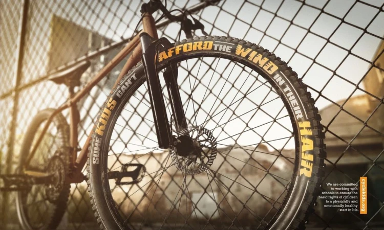 A bicycle is secured to a chain link fence with the words 'SOME KIDS CAN'T AFFORD THE WIND IN THEIR HAIR' written on the tire.
