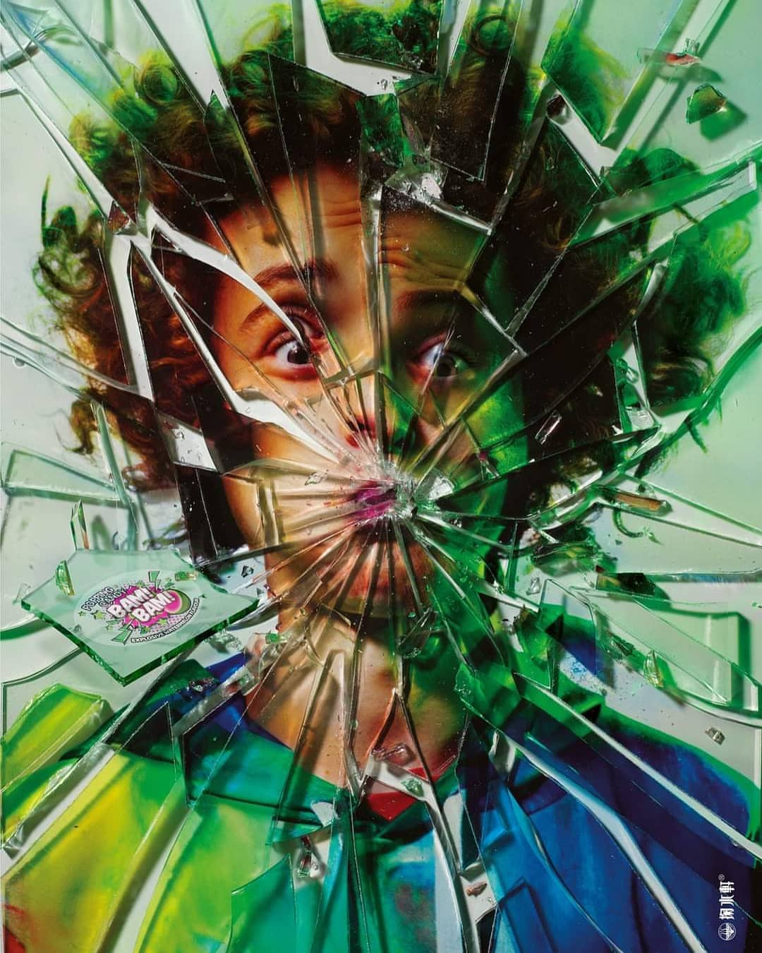 Conceptual portrait of a woman with curly hair shown through a shattered glass surface with reflected colors of blue, green, and yellow.