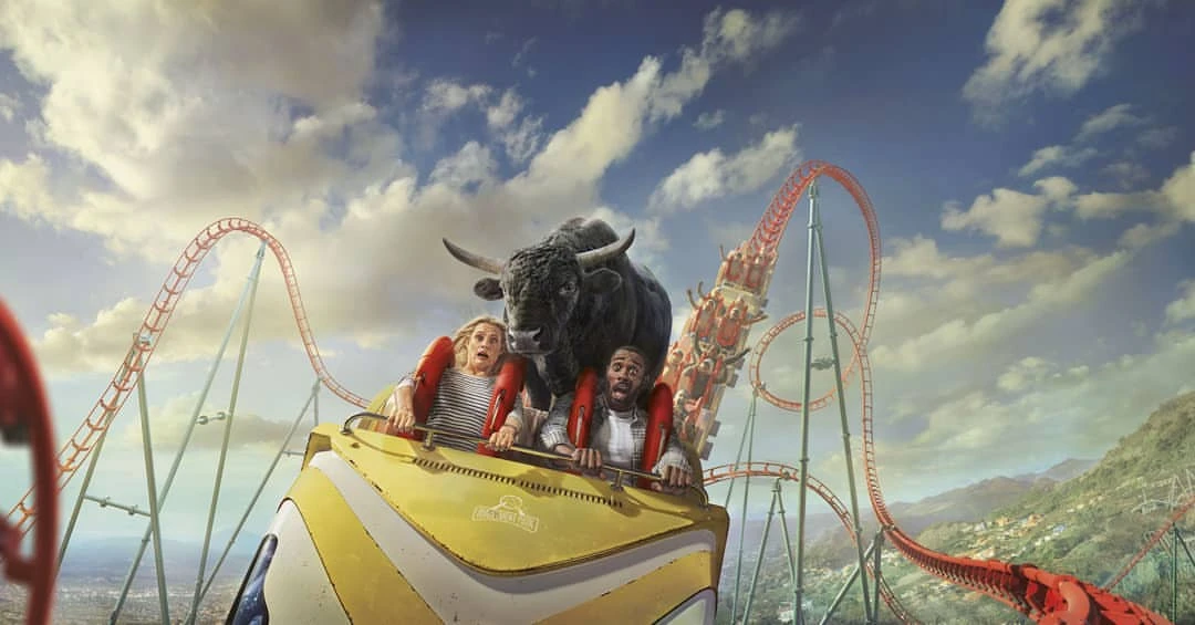 Two people riding a roller coaster with a bull superimposed in front of them, against a backdrop of a blue sky and mountains.