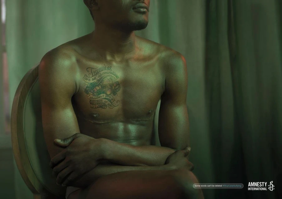 Torso of a shirtless African American male with a tattoo, seated with arms crossed, in a dimly lit indoor setting for Amnesty International.