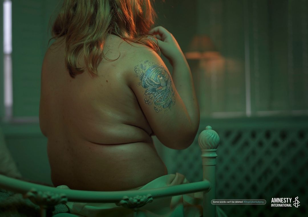 Back view of a woman sitting on a bed in a dark room with a tattoo on her arm, advocating against cyberbullying with an Amnesty International logo.