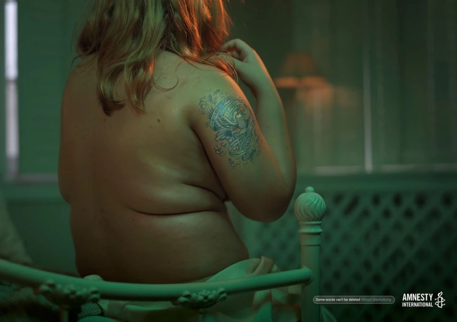 Back view of a woman sitting on a bed in a dark room with a tattoo on her arm, advocating against cyberbullying with an Amnesty International logo.