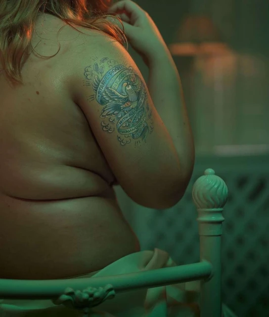 Close-up of an adult female's back and arm featuring a tattoo, set indoors, with a bed frame visible in the foreground.