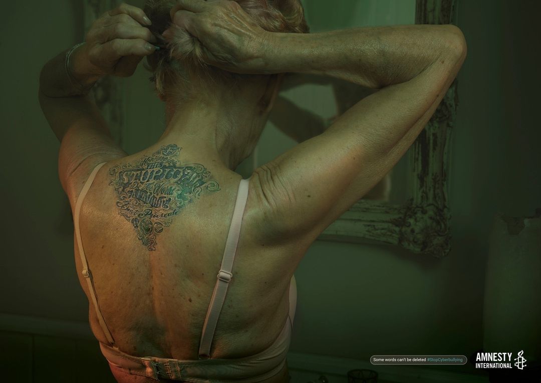 Older woman with tattoo on back raises her arms, tying her hair up. Interior, soft lighting. Amnesty International logo in corner.