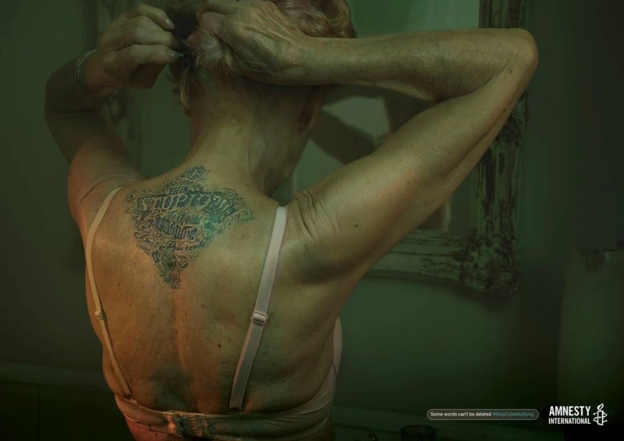 Older woman with tattoo on back raises her arms, tying her hair up. Interior, soft lighting. Amnesty International logo in corner.