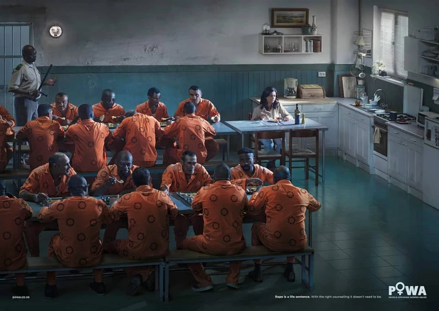 A woman sits at a table in a cafeteria or institutional kitchen setting, surrounded by several male prisoners in orange uniforms, with a guard.