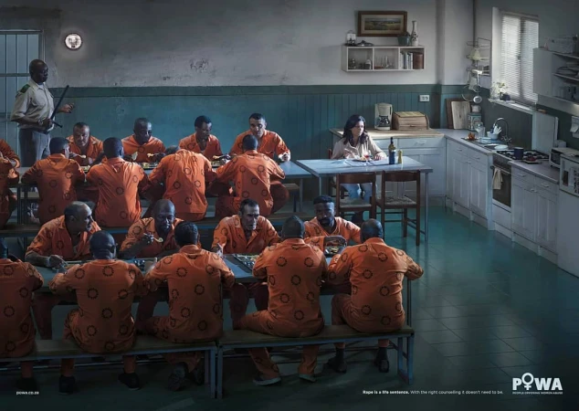 A woman sits at a table in a cafeteria or institutional kitchen setting, surrounded by several male prisoners in orange uniforms, with a guard.
