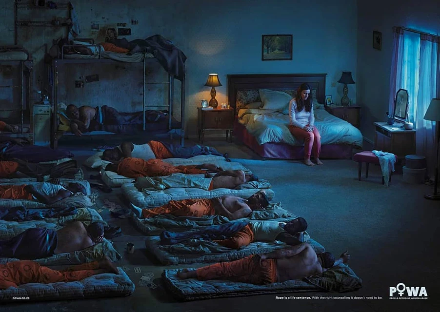 A conceptual photo with a woman sitting on a bed in a room with several men sleeping on floor mattresses and bunkbeds in dim lighting.