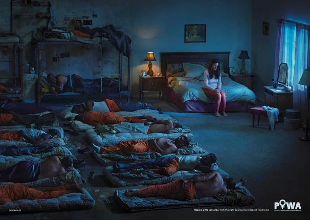 A conceptual photo with a woman sitting on a bed in a room with several men sleeping on floor mattresses and bunkbeds in dim lighting.