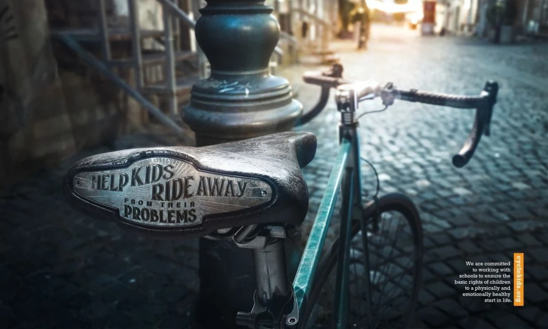 Close-up of bicycle seat with 'Help Kids Ride Away From Their Problems' text against a brick street and city street with buildings.