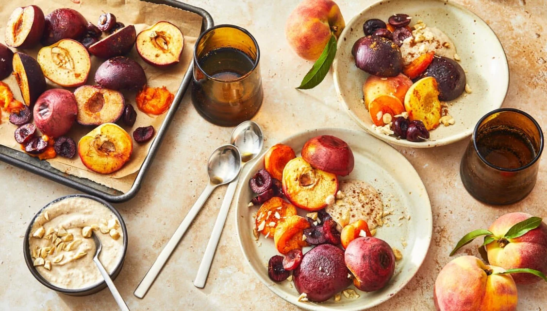 Overhead shot of ripe peaches, plums, and cherries served on plates with sauce, with a baking sheet, bowl, and glasses alongside.