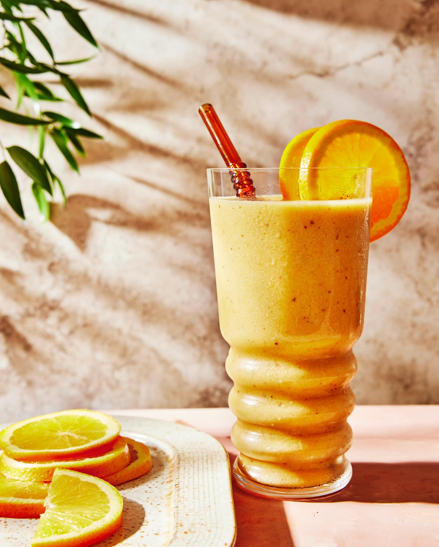 An orange smoothie with orange slices in a tall glass, on a table with plant leaves in the background. Bright and vibrant food photography.
