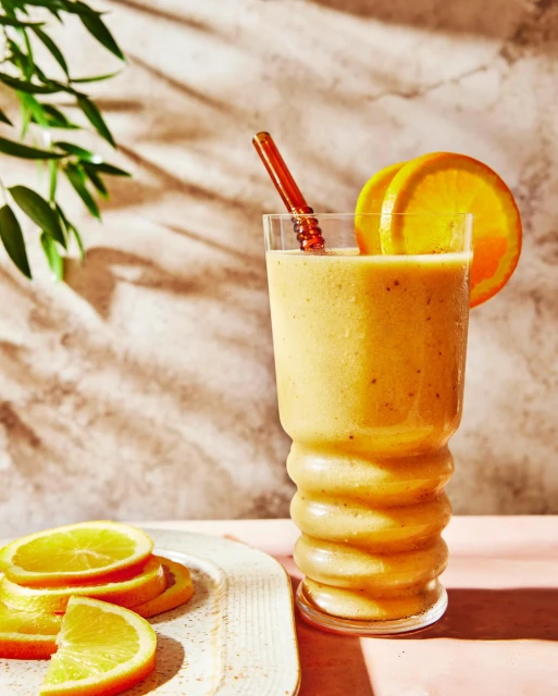 An orange smoothie with orange slices in a tall glass, on a table with plant leaves in the background. Bright and vibrant food photography.