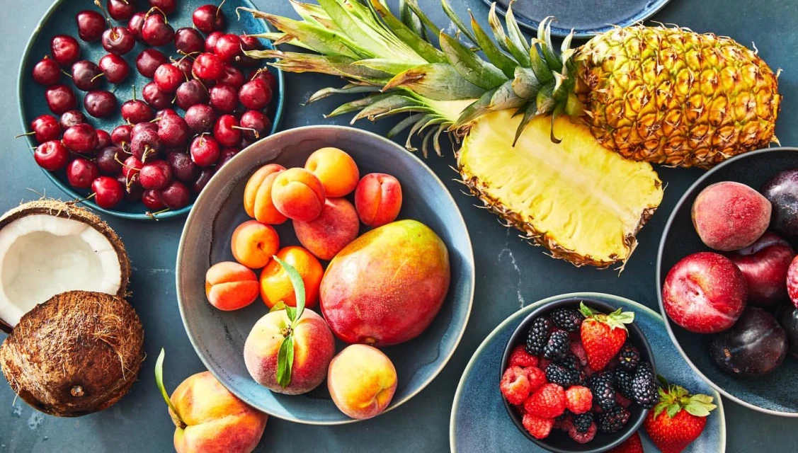Overhead shot of plates and bowls filled with tropical fruits including pineapple, mango, peaches, plums, raspberries, blackberries, cherries, and coconut.