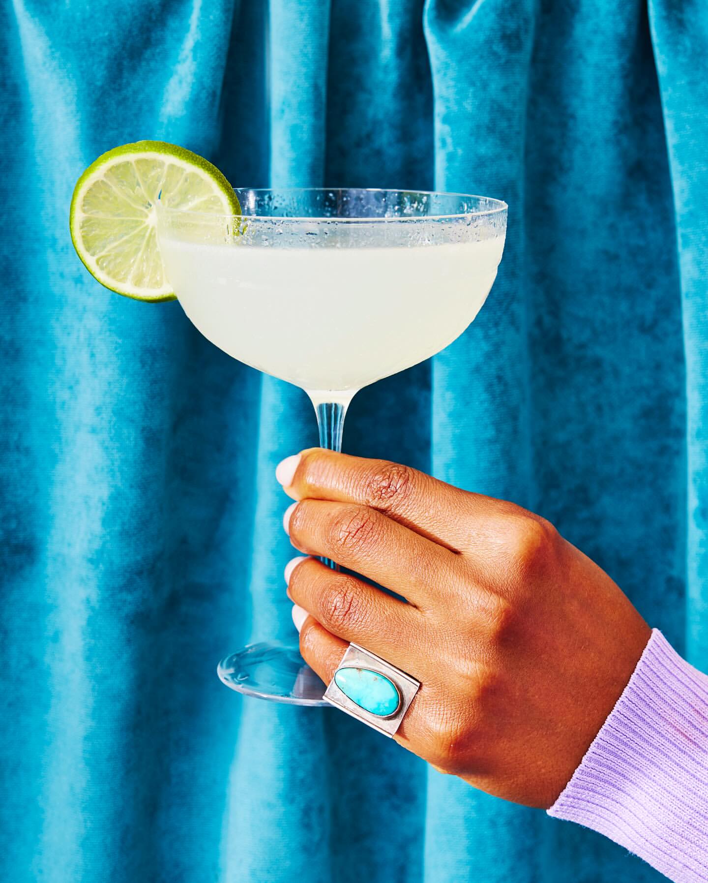 Hand holding a coupe glass filled with a clear cocktail garnished with a lime slice, set against a teal velvet curtain backdrop.