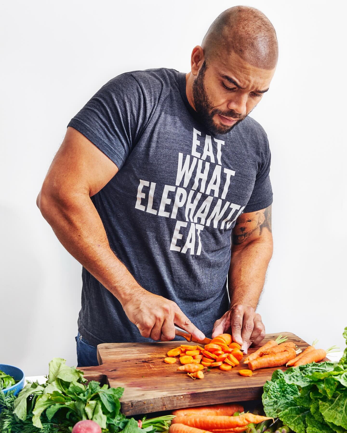 Adult man slicing carrots on a wooden cutting board surrounded by fresh vegetables in a bright kitchen setting, wearing a t-shirt.