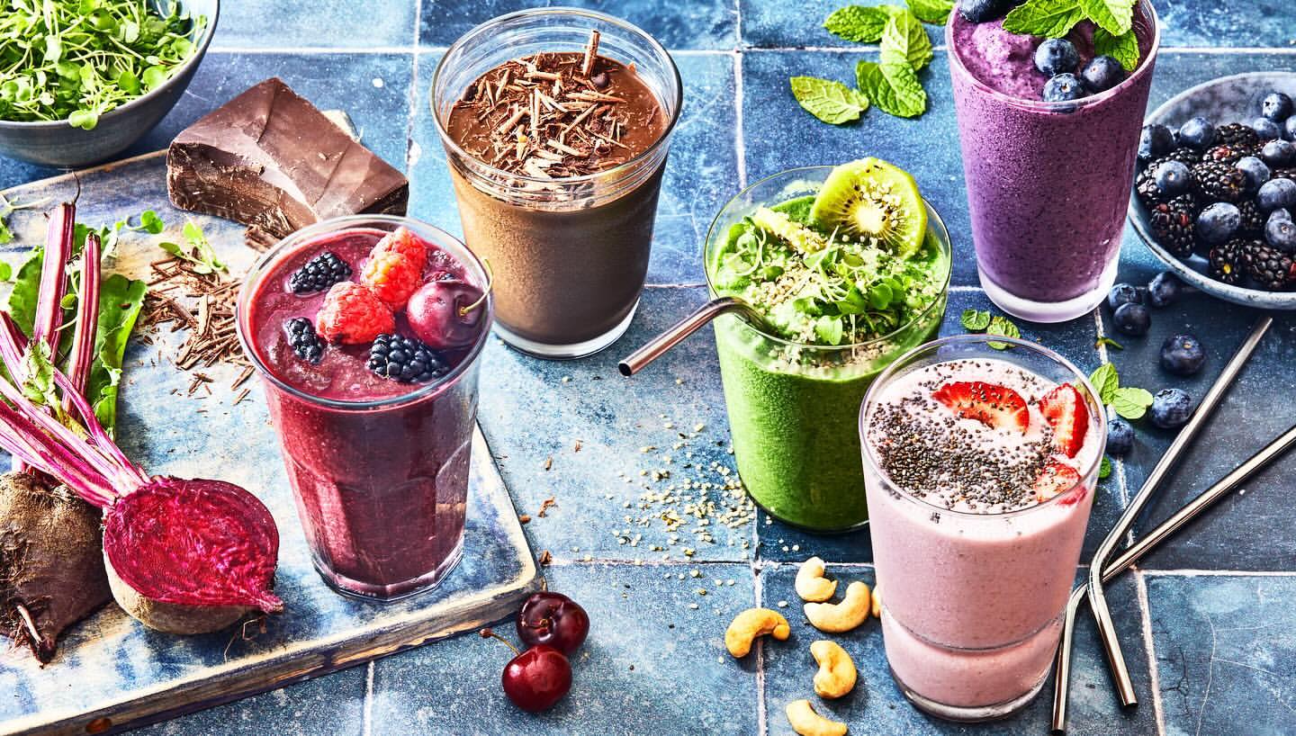 A variety of vibrant, colorful smoothies in glasses, adorned with fruits, nuts, and seeds, displayed on a blue-tiled surface with ingredients.