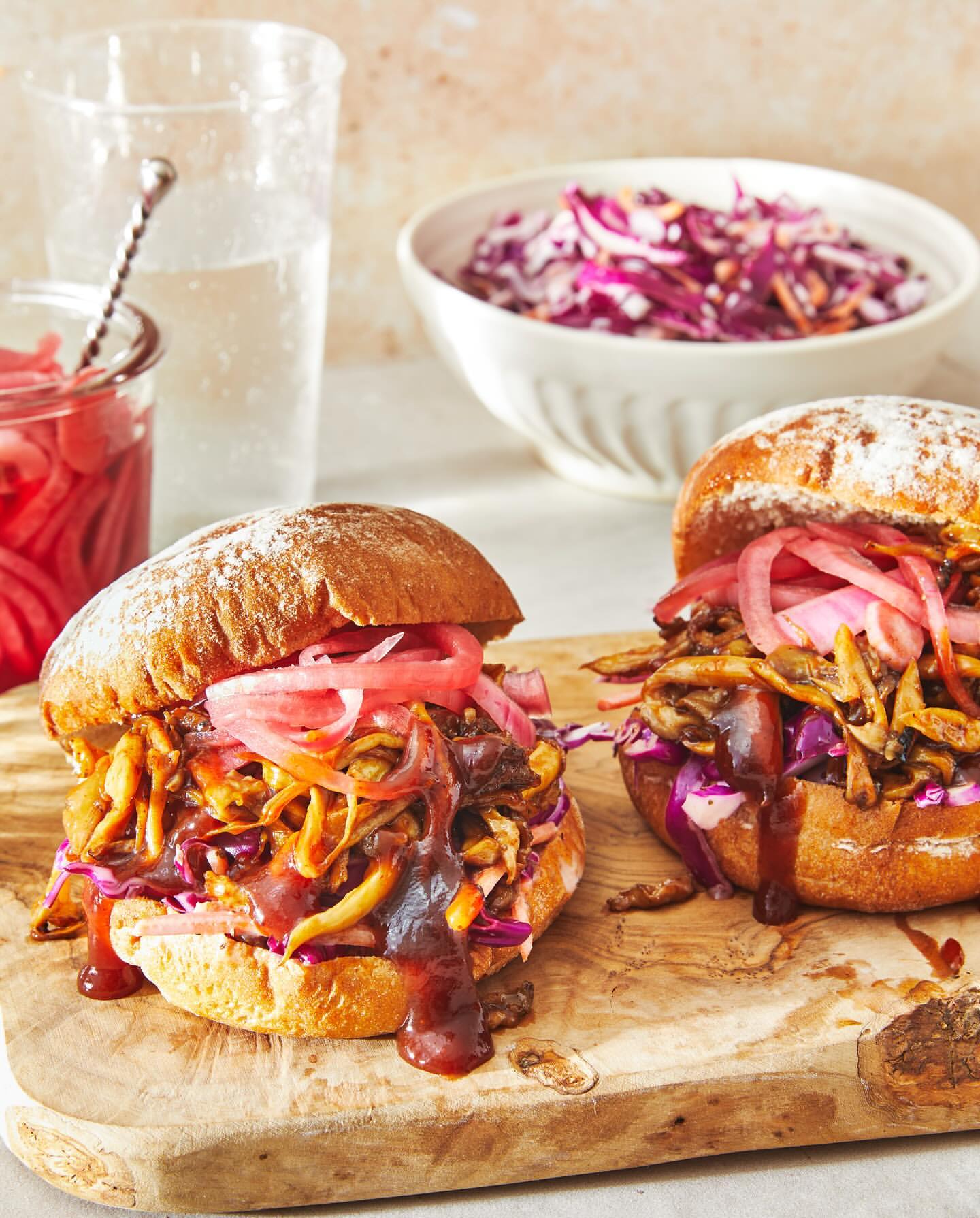 Two sandwiches with pulled meat, pickled onions, and sauce on a wooden board, accompanied by a side of coleslaw and water.