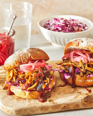 Two sandwiches with pulled meat, pickled onions, and sauce on a wooden board, accompanied by a side of coleslaw and water.