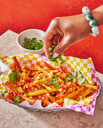 Overhead shot of seasoned sweet potato fries topped with salsa in a checkered basket with a hand adding fresh cilantro.
