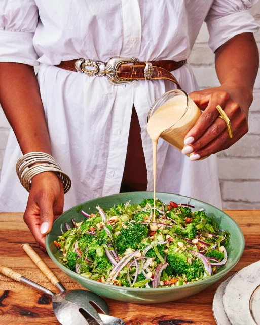 Person pouring dressing on a vibrant broccoli salad on a wooden table with cooking utensils, wearing a white shirt and jewelry.