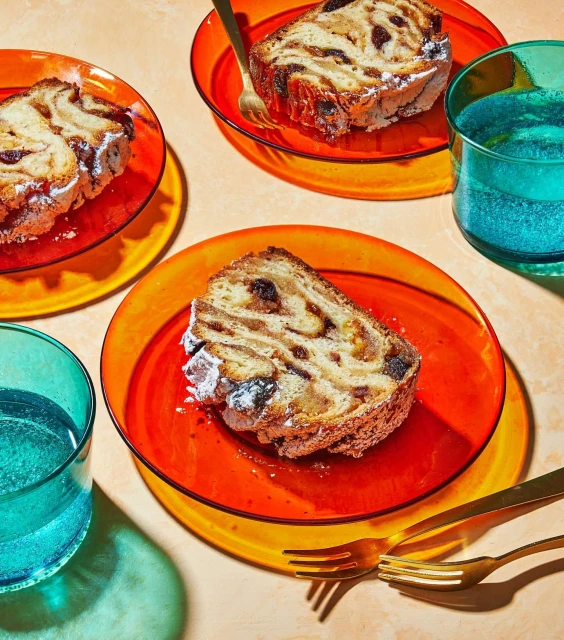 Overhead shot of sliced stollen bread on colorful plates with golden forks, accompanied by glasses of sparkling water.