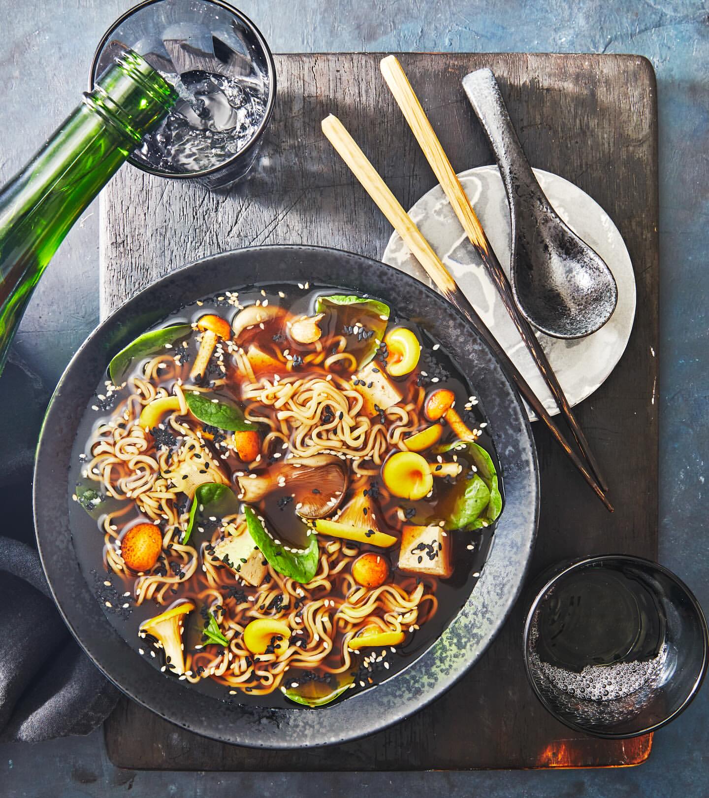 Overhead shot of noodle soup with vegetables and mushrooms being poured from a green bottle, accompanied by chopsticks and a spoon.